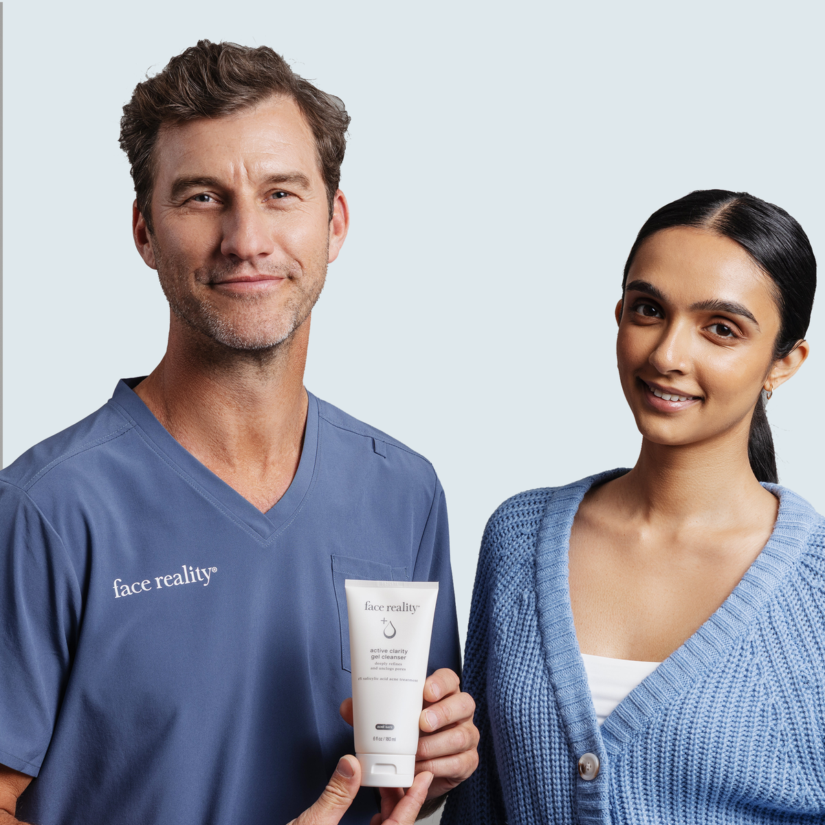 A man in blue Face Reality Skincare scrubs and a woman in a light blue cardigan hold a white tube of Face Reality active clarity gel cleanser together against a light gray studio background.