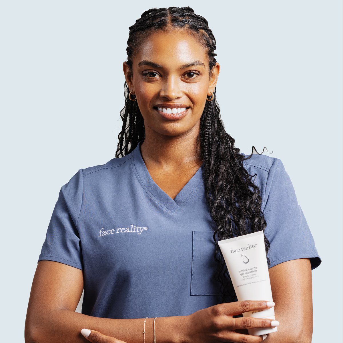 A smiling person in face reality scrubs holding a tube of active clarity gel cleanser against a light gray background